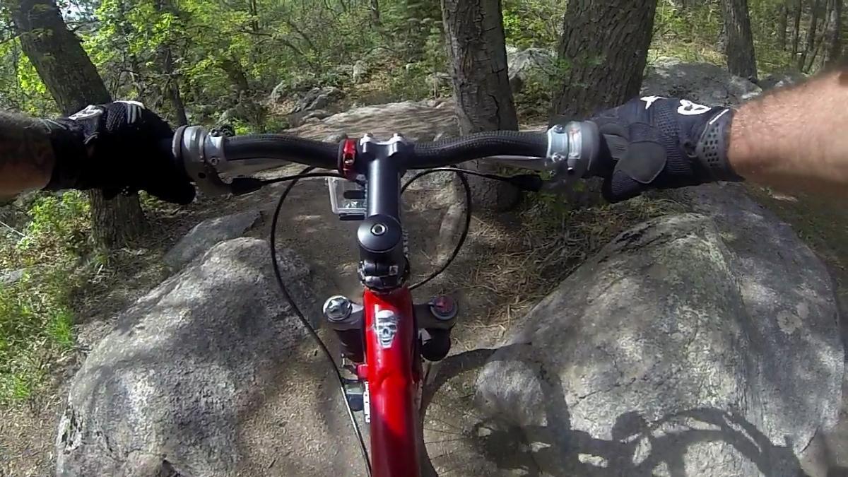 Mountain bike handlebars viewed from the rider's perspective, with hands gripping the grips. The path is rocky, surrounded by trees and greenery, indicating an outdoor trail. The frame is a bold red color, and there are details of the bike's components visible, including brake levers and a digital display. Cheyenne Mountain State Park mountain bike trail.