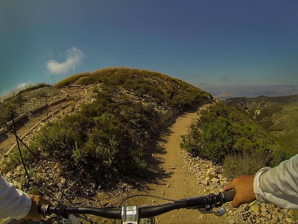 Alt text: A mountain biking perspective showing a dirt trail winding along a hillside with green vegetation and a clear blue sky in the background. The handlebars of the bike are visible in the foreground. Montana De Oro mountain bike trail.