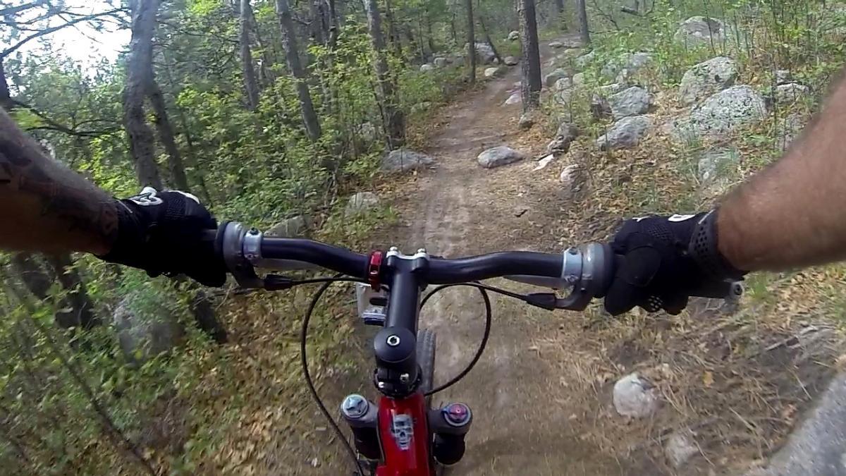 A view from the handlebars of a mountain bike navigating a winding dirt trail surrounded by trees and large rocks. The rider's arms are visible, with one hand gripping the handlebar and the other positioned on the brake, showcasing a focus on the biking experience in a wooded environment. Cheyenne Mountain State Park mountain bike trail.