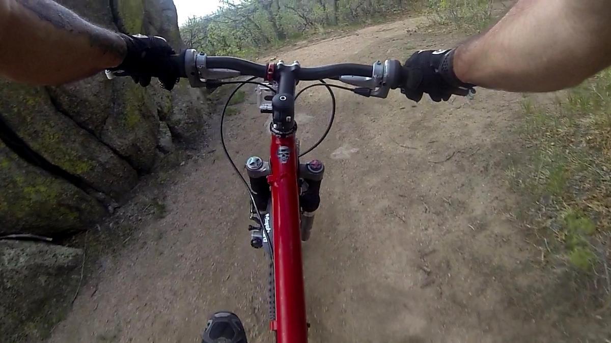 Alt tag: View from the handlebars of a red mountain bike navigating a dirt trail surrounded by rocky terrain and vegetation. Cheyenne Mountain State Park mountain bike trail.