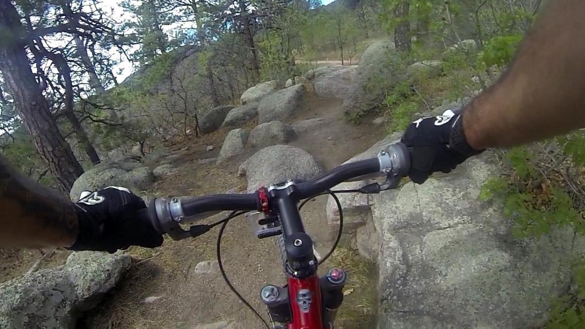 A close-up view of a mountain biker's hands gripping the handlebars of a red bike while navigating a rocky trail surrounded by trees. Cheyenne Mountain State Park mountain bike trail.