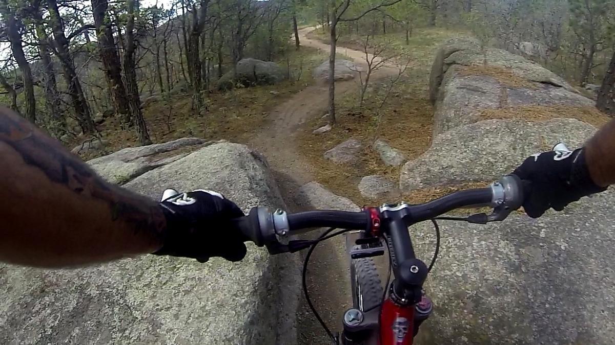 A close-up view from the handlebars of a mountain bike, showing rocky terrain and a winding dirt path through a forested area. The image captures the rider's gloved hands gripping the bike's handlebars, with large boulders and trees in the background. Cheyenne Mountain State Park mountain bike trail.