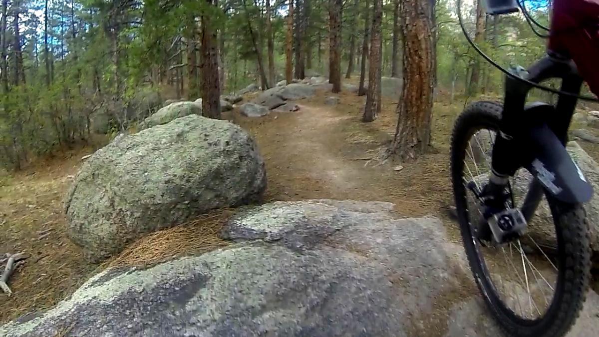 A close-up view of a mountain bike navigating over large rocks on a forest trail. Pine trees and underbrush are visible in the background, creating a natural outdoor setting. The bike's front wheel is prominently displayed, emphasizing the rugged terrain. Cheyenne Mountain State Park mountain bike trail.