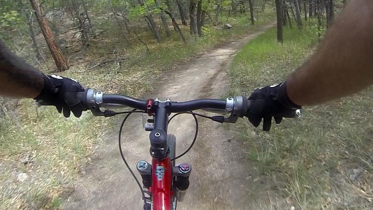 A view from the handlebars of a mountain bike navigating a dirt trail surrounded by trees and greenery. The rider's gloved hands grip the handlebars as the path winds through a forested area. Cheyenne Mountain State Park mountain bike trail.