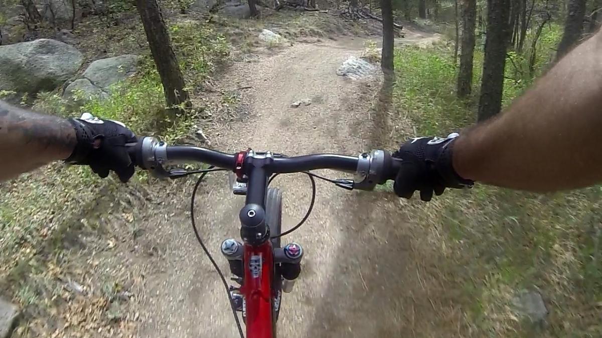 A first-person perspective of mountain biking on a dirt trail surrounded by trees, with hands gripping the handlebars of a red bicycle. The path is narrow and winding, showcasing the natural terrain and lush greenery. Cheyenne Mountain State Park mountain bike trail.
