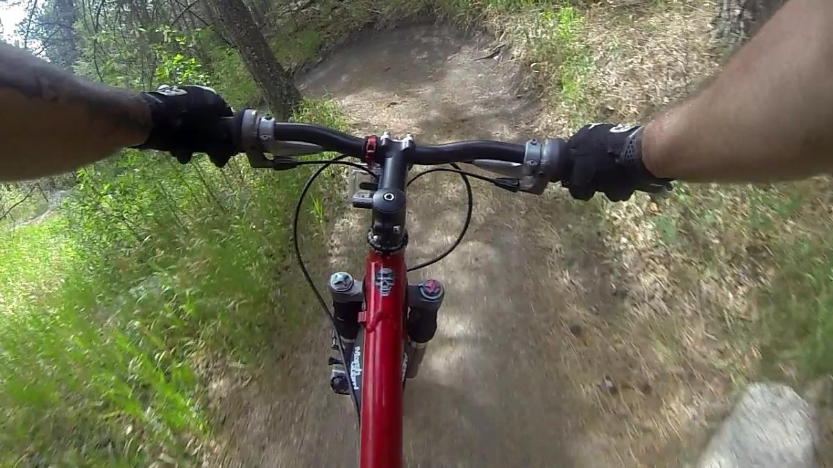 A view from above the handlebars of a mountain bike, showcasing a dirt trail surrounded by green foliage and trees. The rider's gloved hands grip the handlebars as the bike navigates the path. Cheyenne Mountain State Park mountain bike trail.