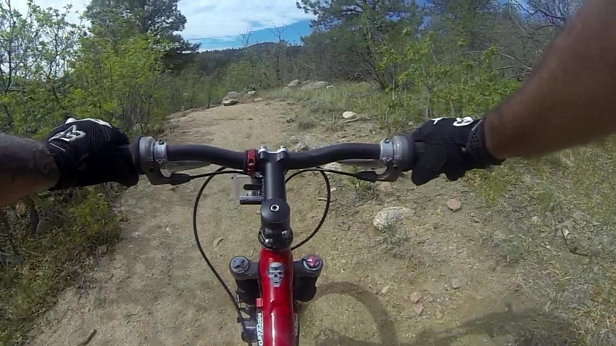 A first-person view of a mountain bike ride on a dirt trail surrounded by greenery, featuring the handlebars and hands of the rider wearing gloves. Cheyenne Mountain State Park mountain bike trail.