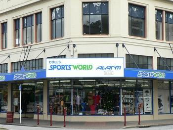 Front view of a retail store named "Sports World," featuring a large sign with the store name and additional branding. The storefront displays various sporting goods and apparel through its windows, with a walkway and colorful signage visible. The building has a classic architectural style with multiple windows.