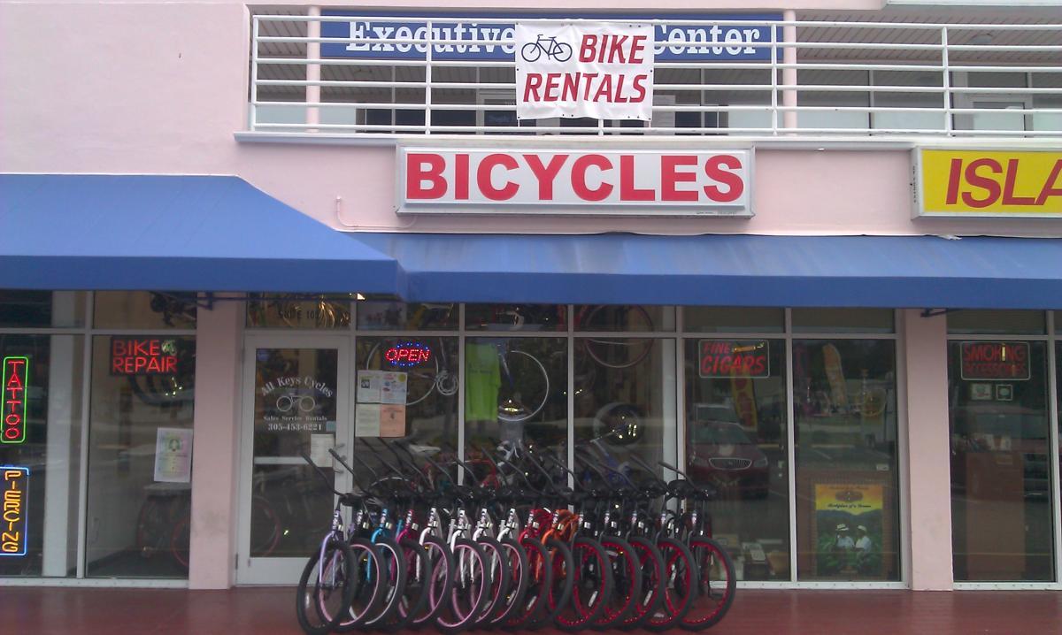 A storefront featuring a "BICYCLES" sign and a banner that says "BIKE RENTALS." The entrance displays a neon "OPEN" sign, and bicycles in various colors are lined up in front. Additional signs on the windows include "BIKE REPAIR," "TATTOO," and "FINE CIGARS." The building has a blue awning and a pink exterior.