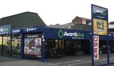A storefront of Avantiplus bicycle shop, featuring a blue and green color scheme, large windows displaying bicycles, and an "OPEN" sign. A banner advertising a bike sale is visible on the right side of the image.