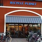 Front view of a bicycle shop named "Bicycle Planet," featuring a large entrance with glass doors and multiple bicycles parked outside. The building has a reddish exterior and a gray awning.