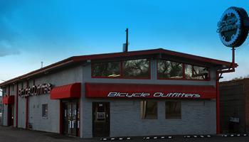 Exterior view of a bicycle shop named "Bicycle Outfitters," featuring a prominent red and gray building with large windows and a circular bicycle gear sign on the roof. The shopfront has a bright red awning displaying the store name. The sky above is blue with some cloud cover.