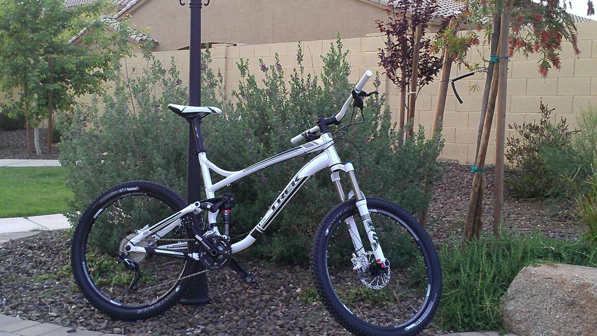 Trek Fuel EX 7: A white mountain bike parked next to a lamp post in a landscaped backyard featuring green shrubs and small trees. The bike has a dual suspension system and thick tires, set against a backdrop of desert landscaping and a tan wall.