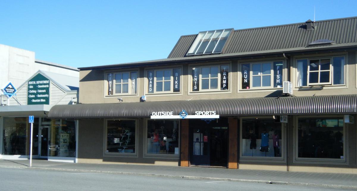 A building facade featuring the sign for "Outside Sports," with large windows displaying outdoor gear. Above, signs indicate various rental services, including skis, snowboards, bikes, and outdoor clothing. The structure has a modern design with a sloped roof and a sunny blue sky in the background.
