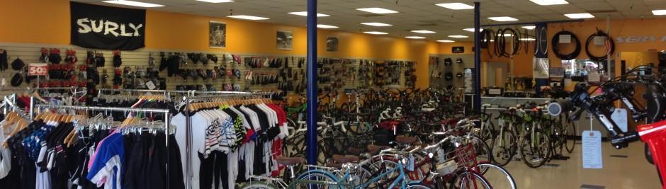Interior view of a bicycle shop featuring a variety of bicycles, cycling apparel, and accessories. The store has a vibrant orange and neutral color scheme, with neatly arranged clothing on racks and numerous bicycles displayed throughout the space. Shelves in the background showcase additional bike accessories.