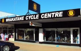 Exterior view of Whakatane Cycle Centre, featuring a black facade with bold white lettering. The shop front showcases several bicycles displayed in large windows, along with a clear blue sky in the background.