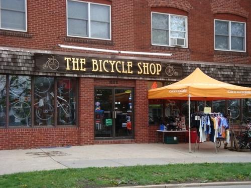 A brick storefront featuring "The Bicycle Shop" sign in gold lettering. The entrance has large windows displaying bicycles inside. Outside, there is a tent with merchandise, including clothing and accessories, on a display table. The setting shows a well-kept sidewalk and grass area in front of the shop.