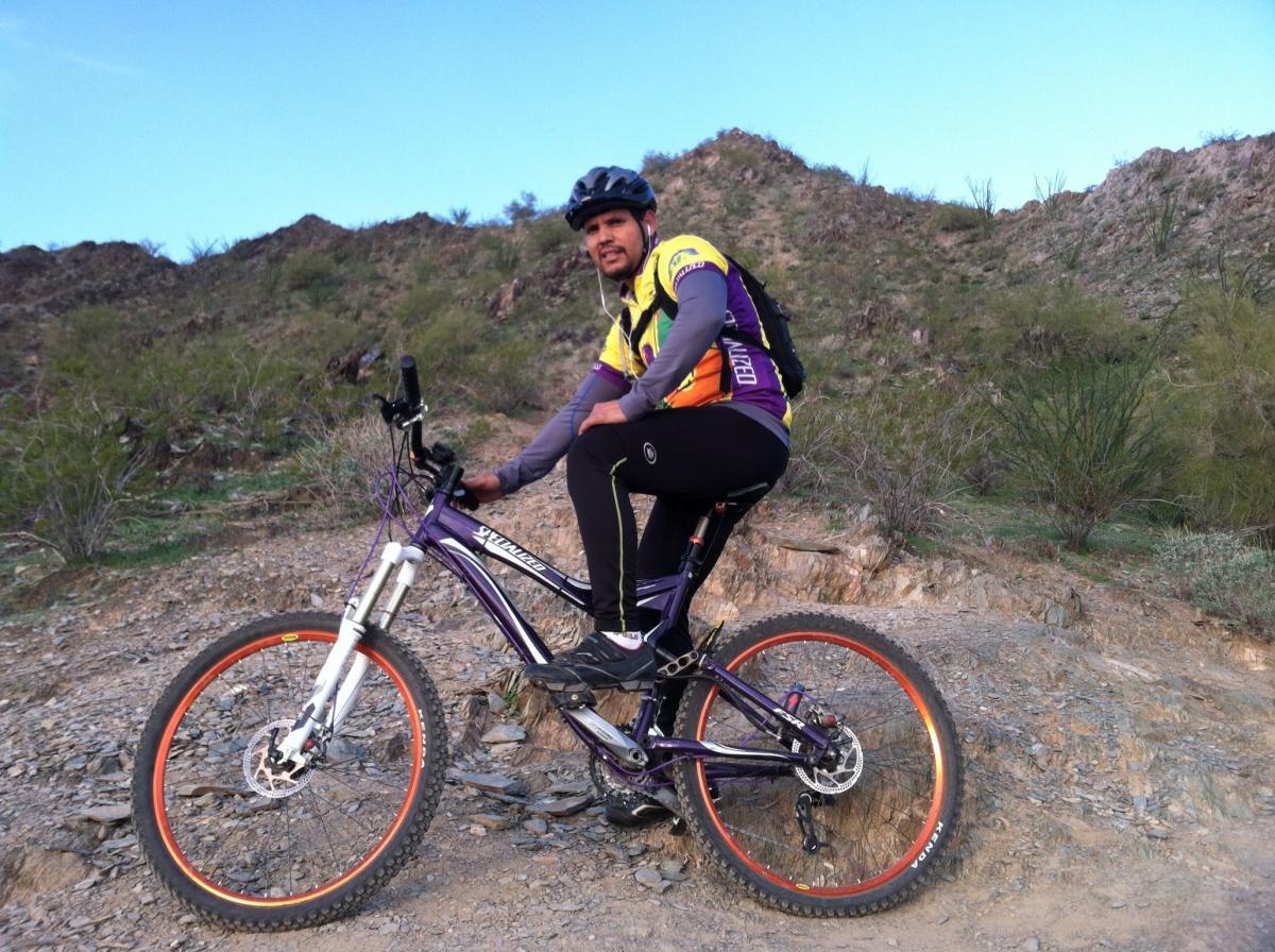 Specialized Stumpjumper FSR Expert: A mountain biker in a colorful cycling jersey and black pants sitting on a purple bike on a rocky trail, surrounded by green shrubs and hills under a clear blue sky.