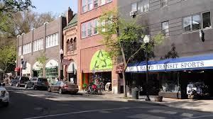 A street view featuring a row of commercial buildings with various storefronts, including a sports shop and a shop with a colorful green sign. Cars are parked along the curb, and trees are visible in the background, indicating a pleasant day.