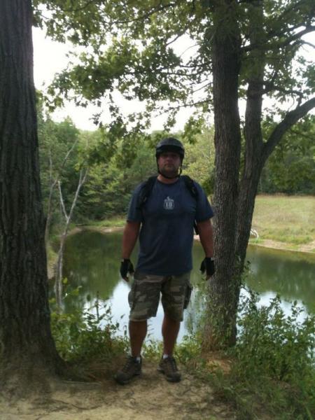 A person wearing a helmet and biking gloves stands near a calm body of water, surrounded by trees and greenery. The individual is dressed in a blue t-shirt and camouflage shorts, positioned on a dirt path with a scenic view of the landscape behind them. Kickapoo mountain bike trail.