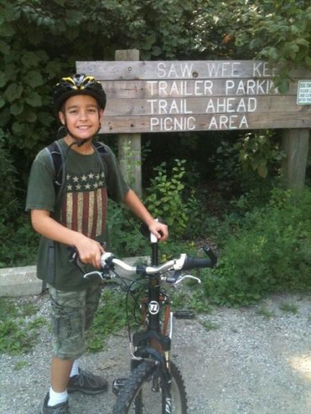 A young boy wearing a bicycle helmet stands next to his bike, smiling in front of a wooden sign that reads "Saw Wee Kee, Trailer Parking, Trail Ahead, Picnic Area." He is dressed in a green t-shirt with an American flag design and camo shorts, surrounded by greenery. Saw Wee Kee Park mountain bike trail.