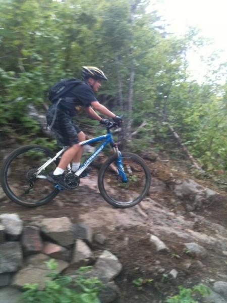 A person riding a blue mountain bike on a rocky trail surrounded by greenery, captured in mid-jump. The rider is wearing a helmet, a black shirt, and shorts, with a backpack. Copper Harbor Trails mountain bike trail.