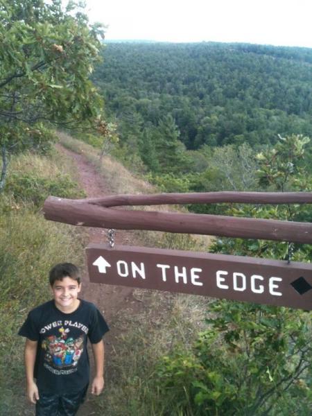 A young boy smiling stands in front of a directional wooden sign that reads "ON THE EDGE," pointing along a trail surrounded by lush greenery and trees. In the background, rolling hills and a scenic view extend into the distance under a cloudy sky. Copper Harbor Trails mountain bike trail.