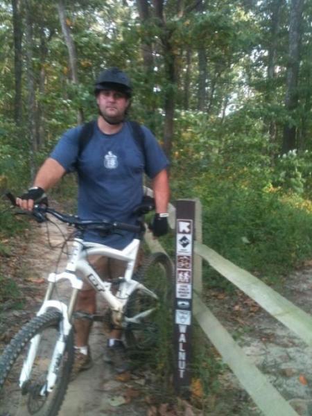 A man wearing a helmet and casual clothing is standing next to a mountain bike on a dirt trail. He is holding the bike's handlebars and looking towards the camera. In the background, a trail marker stands beside a wooden fence, surrounded by greenery and trees, indicating the trail name "Walnut." Brown County Park mountain bike trail.
