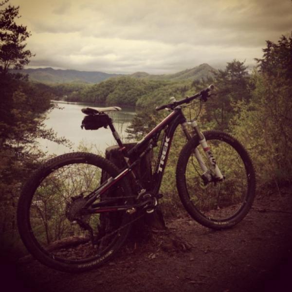 A mountain bike resting against a tree stump, overlooking a serene lake surrounded by green hills and mountains under a cloudy sky. Tsali Recreation Area mountain bike trail.