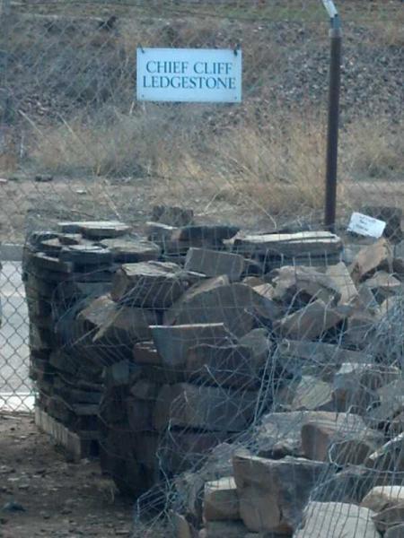 Piles of stacked stones behind a fence, with a sign in the background reading "Chief Cliff Ledgestone." The area is dry with sparse vegetation visible. Cheyenne Mountain State Park mountain bike trail.