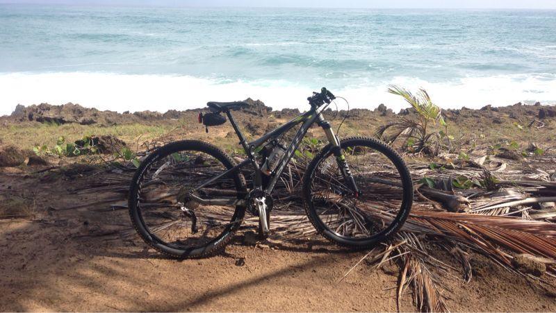 A mountain bike parked on sandy ground near the ocean, surrounded by scattered leaves and rocks. The waves are crashing in the background, with a clear sky above. Cerro Gordo Trail mountain bike trail.