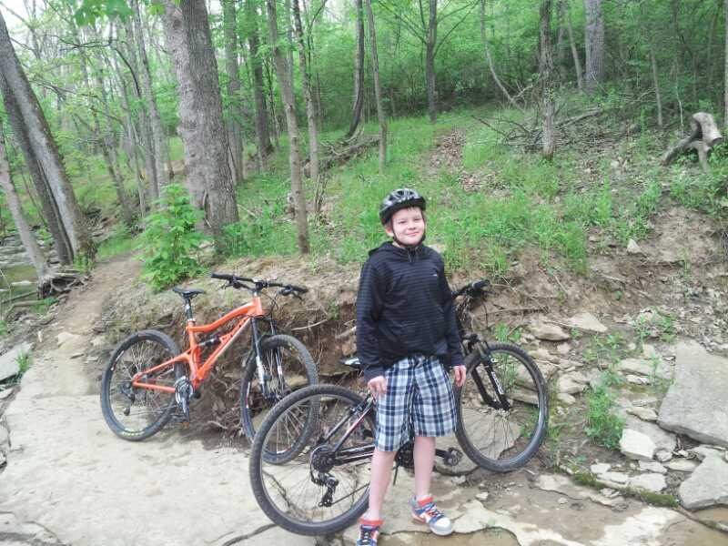 A young boy wearing a black helmet and a black hoodie smiles while standing on a rocky path in a forested area. Two mountain bikes are parked nearby, one orange and the other black, surrounded by green trees and foliage. England Idlewild Mountain Biking Park mountain bike trail.