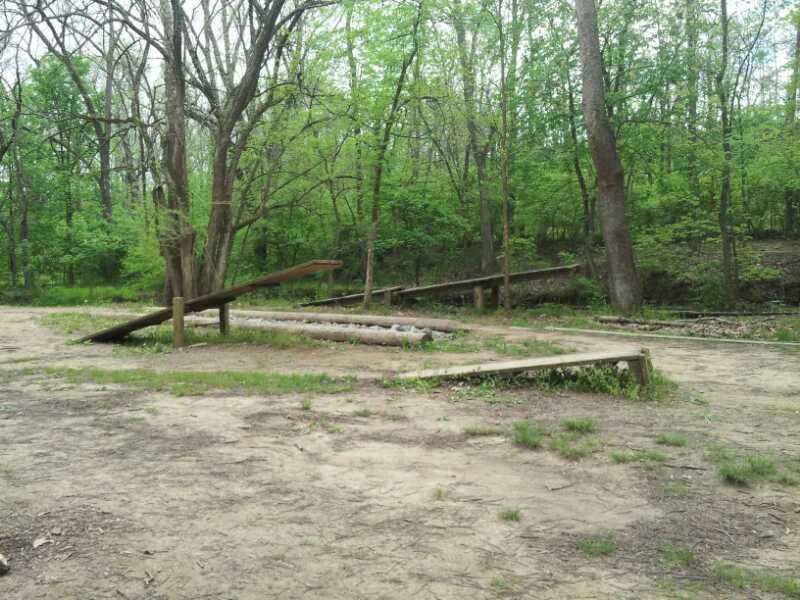 A rustic outdoor scene featuring wooden structures in a forested area. Two wooden ramps are positioned on grassy ground, surrounded by trees and greenery, with a sandy path leading through the space. The setting appears serene and natural, ideal for outdoor activities or relaxation. England Idlewild Mountain Biking Park mountain bike trail.