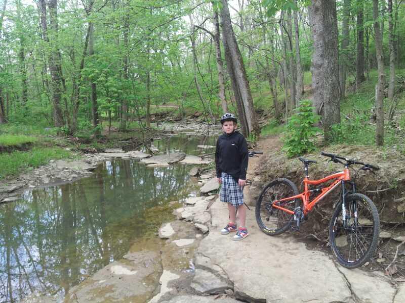 A child wearing a helmet and a black jacket stands by a creek in a lush green forest, next to an orange mountain bike. The setting features rocky terrain and vibrant foliage, creating a serene outdoor atmosphere. England Idlewild Mountain Biking Park mountain bike trail.