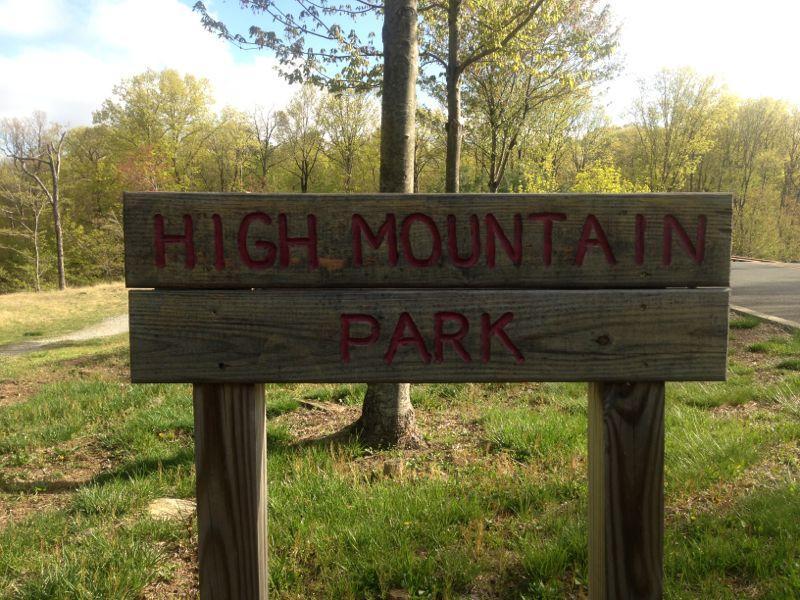 Alt text: A wooden sign that reads "HIGH MOUNTAIN PARK" set in a grassy area surrounded by trees, indicating the entrance to the park. High Mountain Park mountain bike trail.