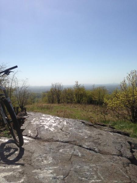 A scenic view from a rocky overlook on a sunny day, featuring a mountain bike positioned on the left side. In the background, rolling hills and trees are visible under a clear blue sky. The foreground shows a large rock surface with some sunlight reflecting off it, surrounded by green grass and sparse shrubs. High Mountain Park mountain bike trail.