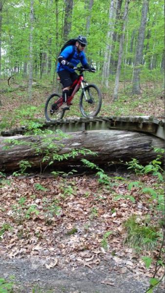 A mountain biker in a blue jacket and black helmet rides over a wooden bridge in a lush green forest, surrounded by trees and fallen leaves. Brown County Park mountain bike trail.