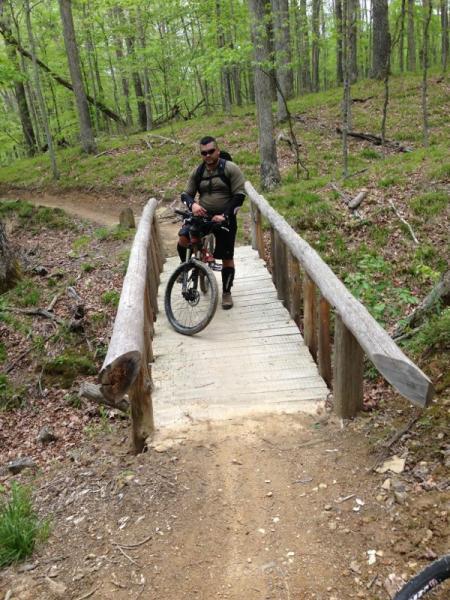 A mountain biker paused on a wooden bridge in a lush, green forest. The trail leads off to the left, with trees and undergrowth surrounding the area. The biker is wearing outdoor gear and is holding onto his bicycle while looking ahead. Brown County Park mountain bike trail.