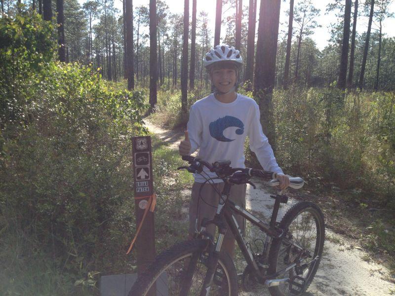 A young boy wearing a helmet and a long-sleeve shirt stands next to his mountain bike at a trailhead in a wooded area. He is smiling and giving a thumbs-up gesture. There are trail signs indicating bike paths, surrounded by tall trees and green vegetation in the background. Bethel Bike Trails mountain bike trail.