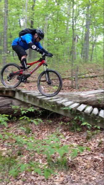 A person wearing a helmet and protective gear rides a mountain bike across a wooden bridge in a lush green forest. The cyclist is focused and balanced as they navigate the uneven surface of the bridge. Surrounding foliage includes trees and underbrush, creating a vibrant, natural setting. Brown County Park mountain bike trail.
