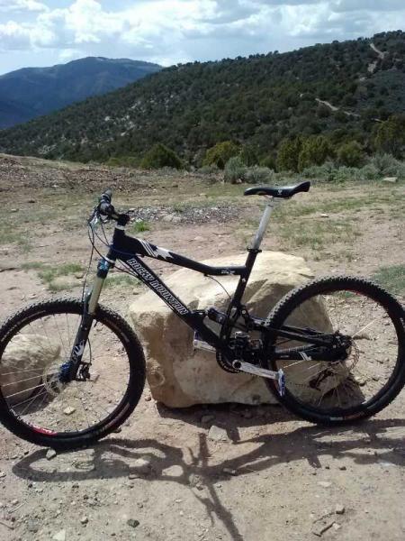 A black mountain bike parked next to a large rock, set against a backdrop of rolling hills and a cloudy sky. The bike features thick tires and a sturdy frame, indicating it's designed for off-road trails. Berry Creek Loop mountain bike trail.