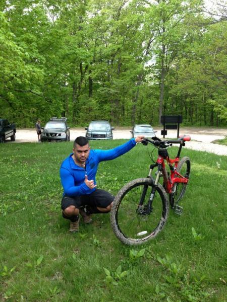 A person in a blue long-sleeve shirt squatting next to a red mountain bike on grass, giving a thumbs-up. In the background, several parked cars and trees can be seen, indicating an outdoor setting. Brown County Park mountain bike trail.