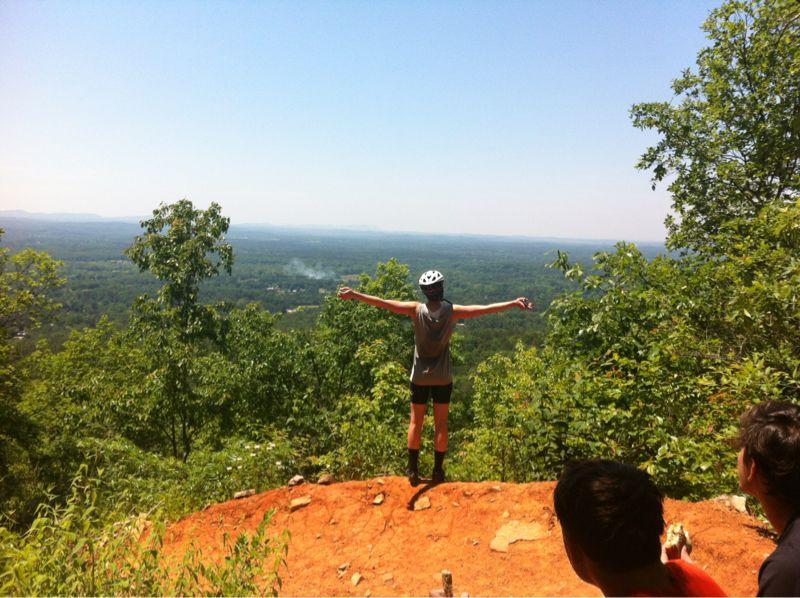 A person wearing a helmet stands on the edge of a rocky outcrop, arms outstretched, overlooking a vast green landscape under a clear blue sky. Lush trees surround the area, while a few other individuals are partially visible in the foreground, observing the view. Coldwater Mountain mountain bike trail.