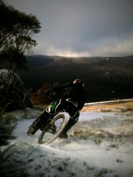 A mountain biker navigating a snowy trail, leaning into a turn surrounded by a scenic mountainous landscape. The scene conveys a sense of adventure and outdoor activity in winter conditions. Cannonball mountain bike trail.