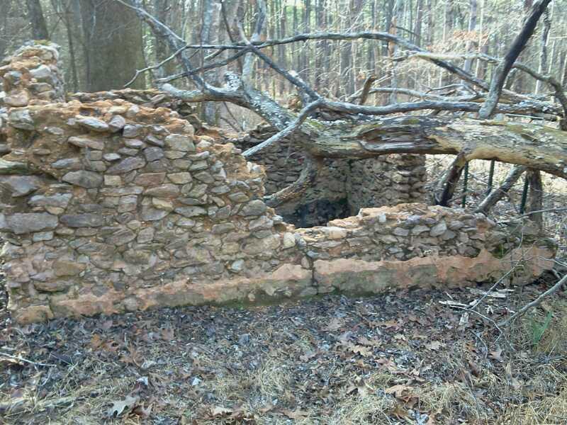 Alt text: Ruins of a stone structure partially covered by fallen branches and surrounded by wooded terrain with dried leaves on the ground. Buncombe - Brickhouse Recreation Area mountain bike trail.