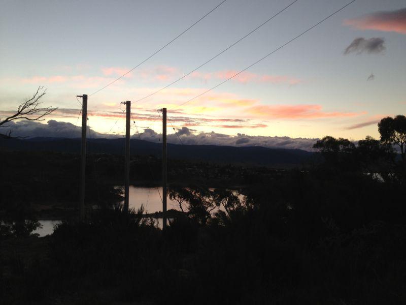 Sunset view over a landscape featuring utility poles, mountains in the background, and a calm body of water reflecting the sky. Fluffy clouds are illuminated with hues of pink and orange as daylight fades. She