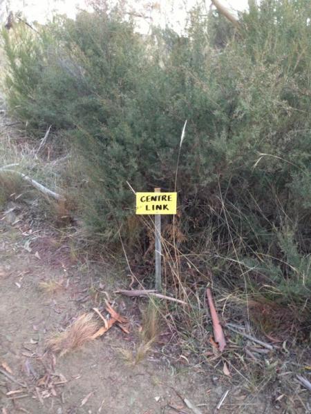 A small yellow sign reading "CENTRE LINK" mounted on a wooden stake, set against a backdrop of green bushes and dry grass in a natural outdoor environment. Centre link mountain bike trail.