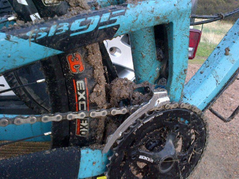 Close-up of a mountain bike frame covered in mud, showcasing the chain, derailleur, and tire. The bike features a blue color scheme and appears to have been used in off-road conditions. Wildcat Mountain / Monarch mountain bike trail.