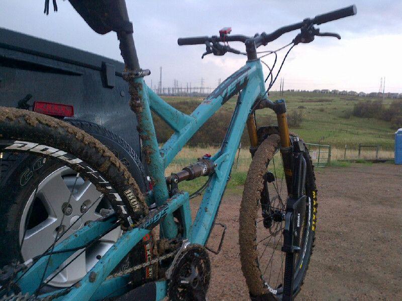 A muddy mountain bike is parked next to a vehicle, showcasing its turquoise frame and dirt-covered wheels. The background features an open landscape with grass, distant power lines, and a cloudy sky. Wildcat Mountain / Monarch mountain bike trail.