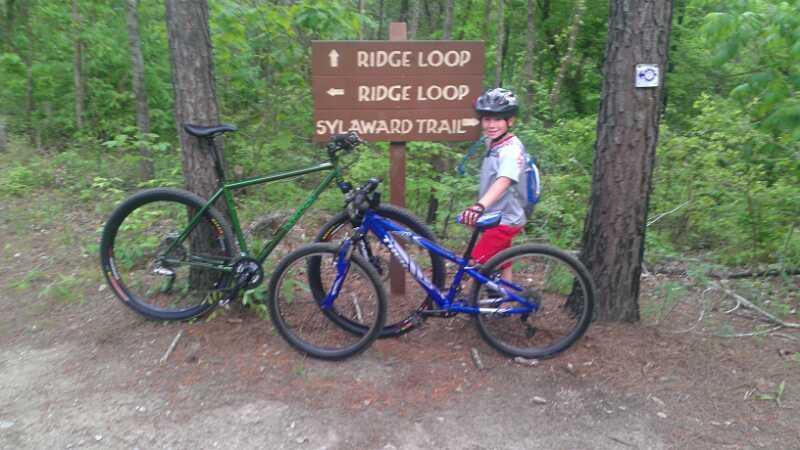 A young boy wearing a helmet is smiling beside two mountain bikes, parked near a wooden trail sign. The sign indicates the directions for "Ridge Loop" and "Sylaward Trail," surrounded by lush green trees in a natural outdoor setting. Sylaward mountain bike trail.